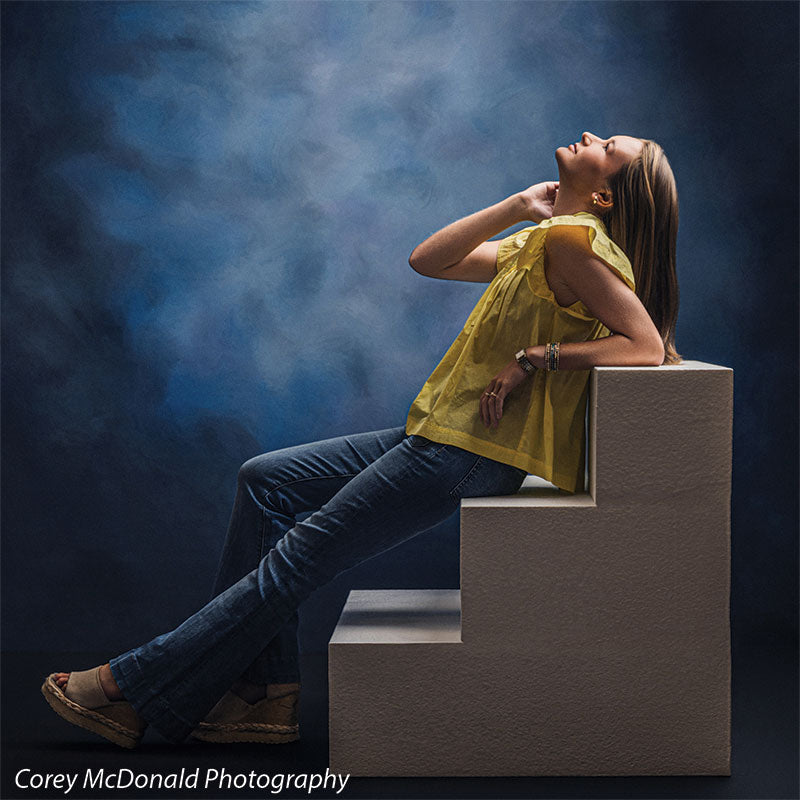 Neo Classical blue mottled texture photography backdrop shown with a senior girl in a yellow ruffle-sleeve top and blue flared jeans reclining against the white foam steps looking upward on white foam posing steps OM-1500