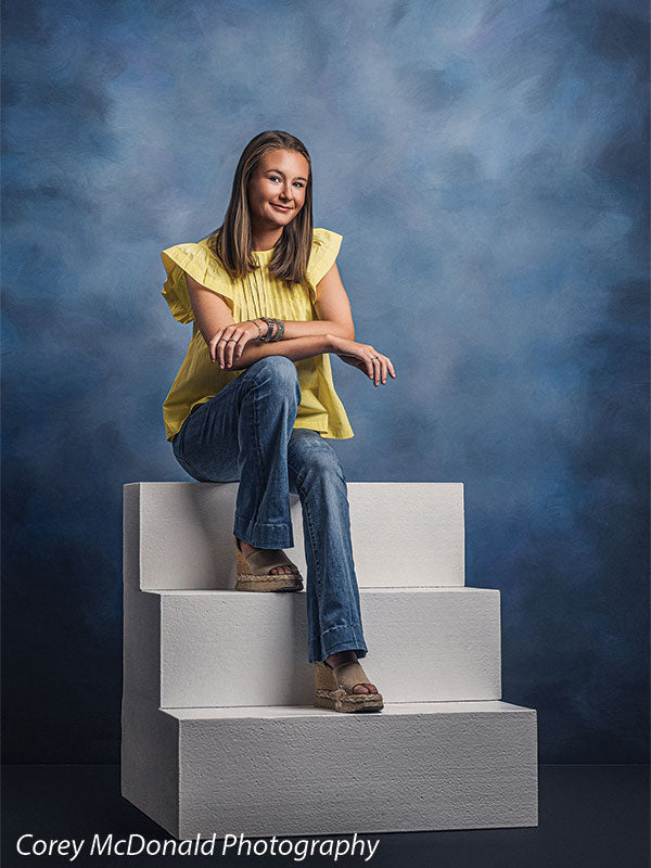 Neo Classical blue mottled texture photography backdrop shown with a senior girl in a yellow ruffle-sleeve top and blue flared jeans seated on the white foam steps with one arm resting on her knee on white foam posing steps OM-1500
