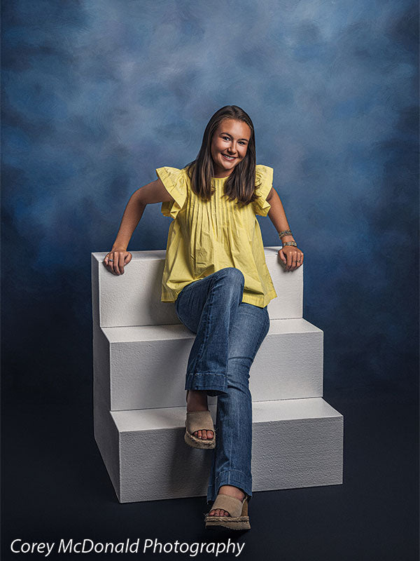 Neo Classical blue mottled texture photography backdrop shown with a senior girl in a yellow ruffle-sleeve top and blue flared jeans seated on the white foam steps leaning back on her hands on white foam posing steps OM-1500