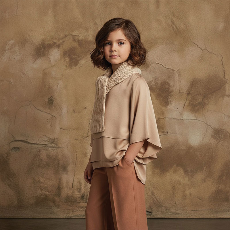 Young girl with wavy brown hair wearing a beige layered top, knit scarf, and brown pants stands slightly angled with hands in pockets against a neutral beige aged plaster texture backdrop.