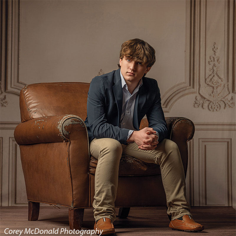Teen boy with wavy light brown hair wearing a blue blazer and khaki pants sits slightly forward in a vintage brown leather chair with hands clasped, set against an elegant beige backdrop with ornate wall panel design.