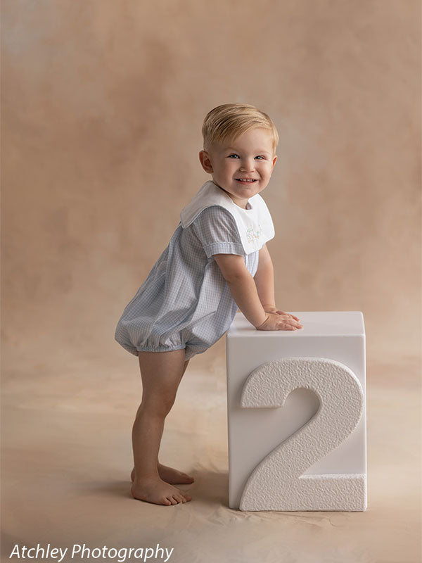A toddler wearing a light blue romper stands barefoot while leaning on a white cube with the number two, smiling toward the camera, posed against a neutral beige portrait studio backdrop.
