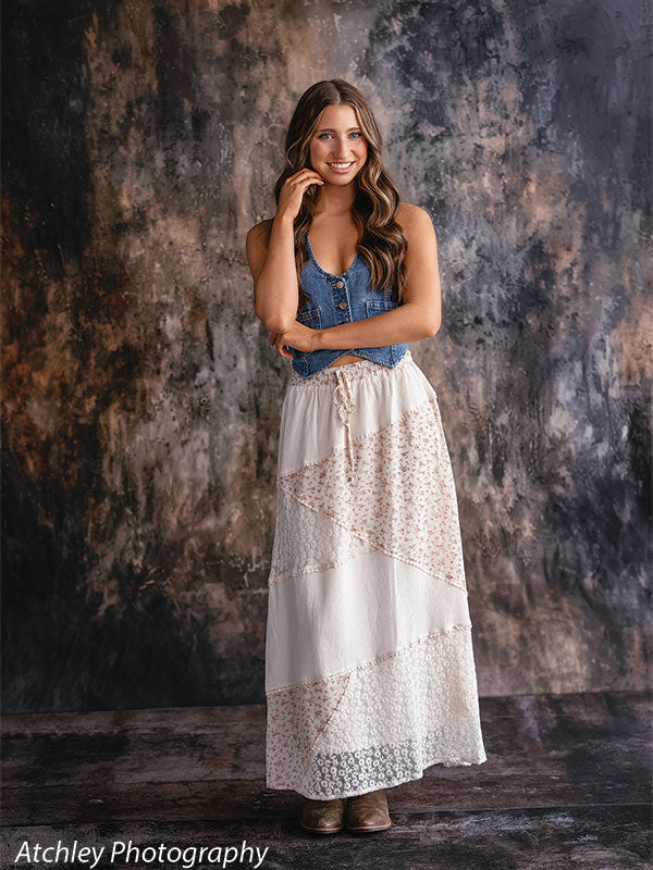 Young woman in a white lace maxi skirt and denim vest standing with one hand near her face, smiling against a neutral brown abstract studio wall backdrop with soft gray and warm earth tone textures.