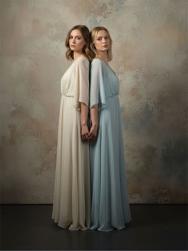 Two women wearing flowing neutral-toned dresses stand back to back holding hands, posed against a softly textured studio backdrop with blended beige and gray painterly tones.
