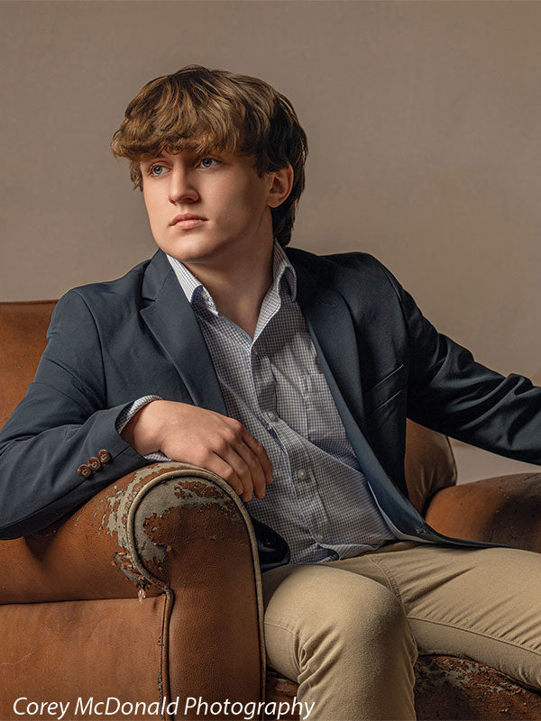 Teen boy with wavy light brown hair wearing a blue blazer and khaki pants sits in a worn leather chair looking off to the side, set against a soft beige vintage wall backdrop with ornate texture and panel detailing.