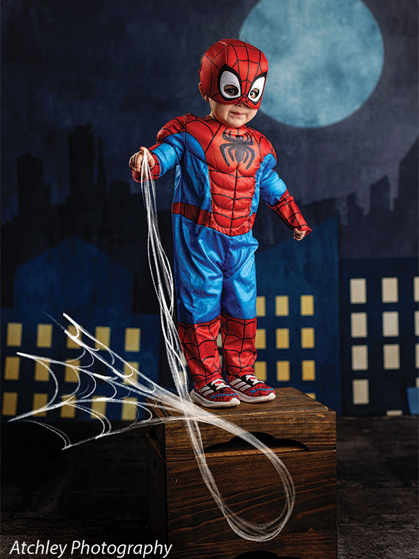 A toddler dressed in a red and blue superhero costume with a matching mask stands on a wooden box holding a web prop, set against a nighttime city skyline backdrop with illuminated windows and a moon.