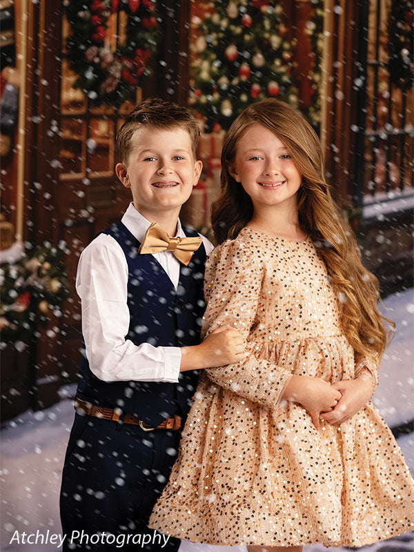 Noel Street snowy Christmas village toy shop photography backdrop shown with a boy and girl standing together as snow falls on the village street CP-8890
