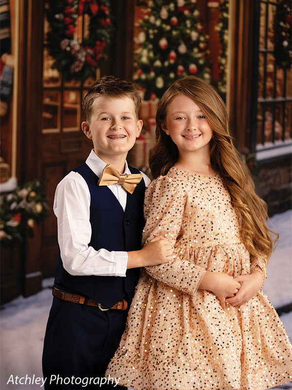 Noel Street snowy Christmas village toy shop photography backdrop shown with a boy in a navy vest and a girl in a gold dress standing together in the snow CP-8890