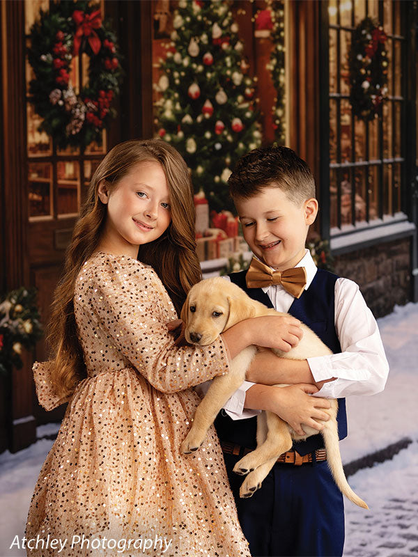 Noel Street snowy Christmas village toy shop photography backdrop shown with a boy and girl holding a yellow Labrador puppy outside the toy shop CP-8890