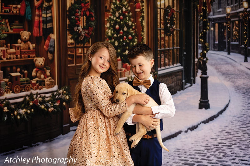 Noel Street snowy Christmas village toy shop photography backdrop shown with a boy and girl holding a yellow Labrador puppy outside the toy shop, wide view CP-8890