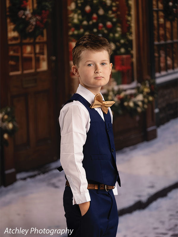 Noel Street snowy Christmas village toy shop photography backdrop shown with a boy in a navy vest and gold bow tie standing on the snowy street CP-8890