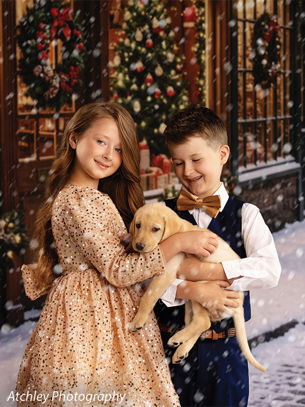 Noel Street snowy Christmas village toy shop photography backdrop shown with two children holding a yellow puppy outside the snowy toy shop CP-8890