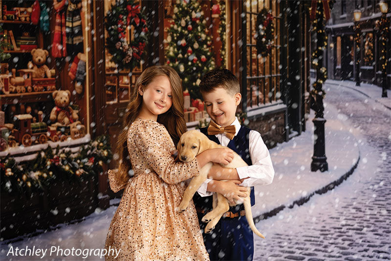 Noel Street snowy Christmas village toy shop photography backdrop shown with two children holding a puppy in front of the lit toy shop, wide view CP-8890