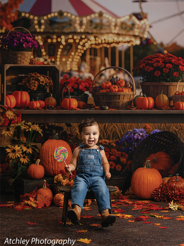 Toddler sitting among large pumpkins and colorful autumn flowers while holding a lollipop, photographed in front of a nostalgic carousel fairground backdrop with harvest baskets and warm seasonal lighting.