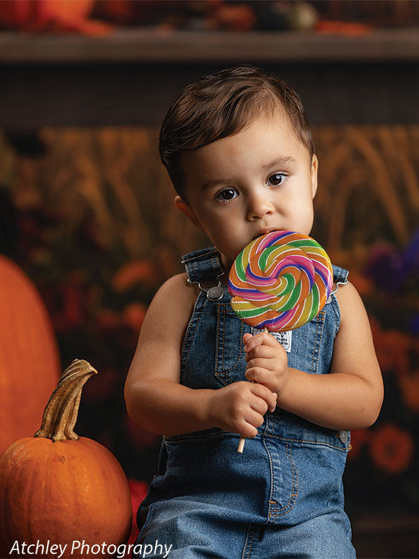 Toddler wearing blue denim overalls sitting beside large orange pumpkins and holding a colorful swirl lollipop, posed in front of a nostalgic fairground backdrop featuring a carousel with glowing lights, baskets of pumpkins, fall flowers, and autumn leaves.