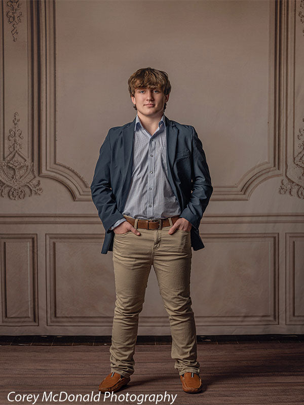 Teen boy with wavy light brown hair wearing a blue blazer, light blue button-down shirt, and khaki pants stands with hands in pockets in front of a neutral beige backdrop featuring ornate vintage wall panel molding and decorative trim.