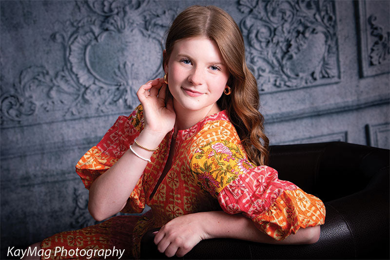 Smiling teen girl in a vibrant boho-style dress leaning on a brown leather chair in front of an ornate textured gray wall photo backdrop, perfect for elegant indoor portraits.