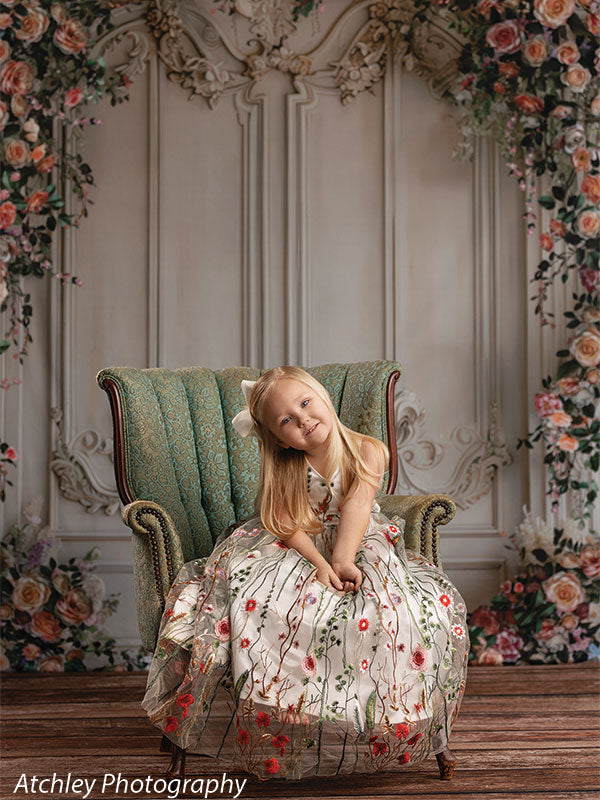 A young girl in a long floral dress sits on a green upholstered chair, tilting her head with a gentle smile, set against an ornate panel wall backdrop surrounded by floral arrangements.
