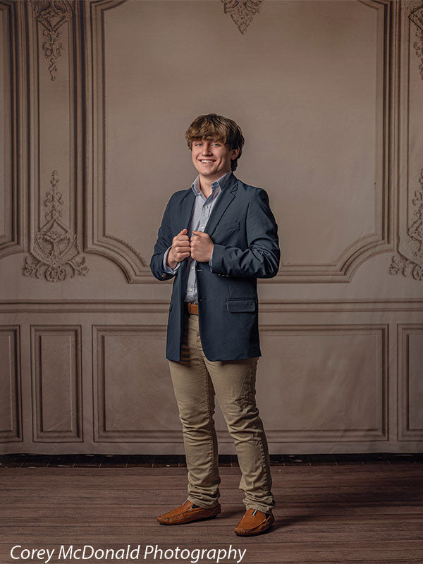 Teen boy wearing a navy blazer, light blue shirt, khaki pants, and brown shoes stands smiling while holding his jacket lapels in front of a beige ornate wall panel backdrop with decorative vintage interior detailing.