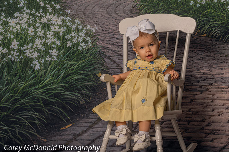 Baby girl wearing a yellow dress with embroidered blue flowers and a white bow headband sits in a white wooden chair on a winding brick garden path backdrop with dense floral borders and greenery.