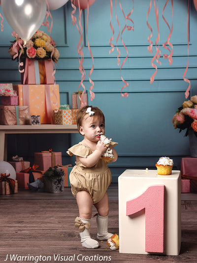 Adorable girl eating cupcake with pink number one birthday block in a balloon-filled, pastel-colored celebration setup.