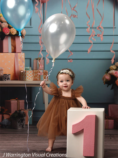 Baby girl holding balloons while standing by a pink number one block, surrounded by presents and colorful birthday balloons.