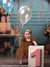 Baby girl holding balloons while standing by a pink number one block, surrounded by presents and colorful birthday balloons.