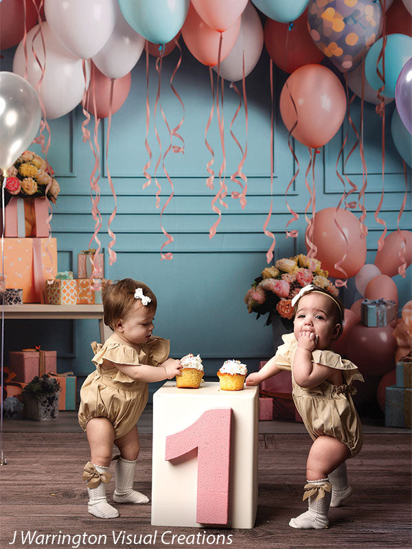 Two baby girls enjoying cupcakes in front of festive Peach Balloon Bliss Cake Smash Backdrop with balloons and flowers.