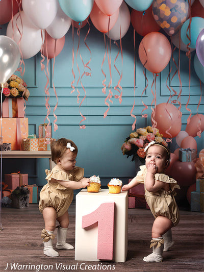 Two baby girls enjoying cupcakes in front of festive Peach Balloon Bliss Cake Smash Backdrop with balloons and flowers.