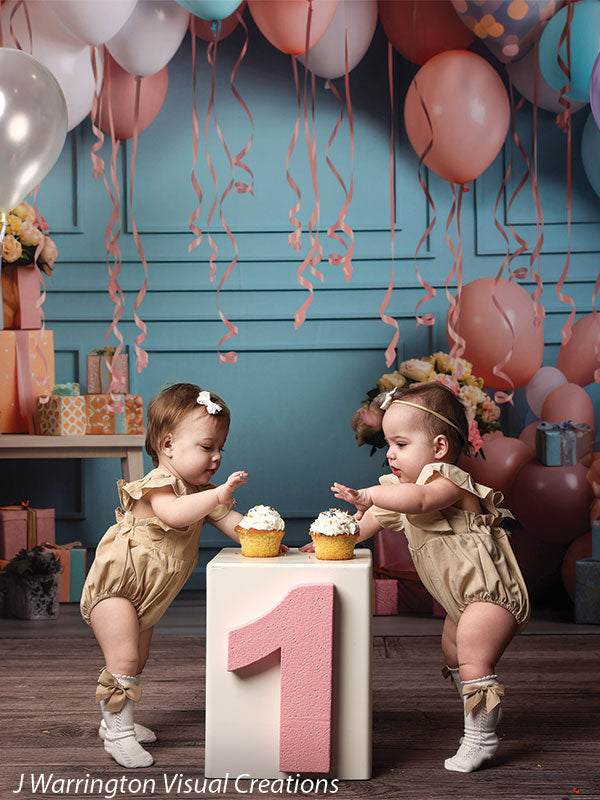 Twin girls with cupcakes celebrating first birthday surrounded by pastel balloons and wrapped gifts.