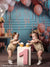 Twin girls with cupcakes celebrating first birthday surrounded by pastel balloons and wrapped gifts.