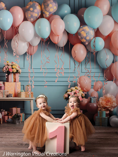 Twin baby girls smiling and standing by pink number one birthday block under peach balloon and ribbon party setup.