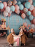 Twin baby girls smiling and standing by pink number one birthday block under peach balloon and ribbon party setup.