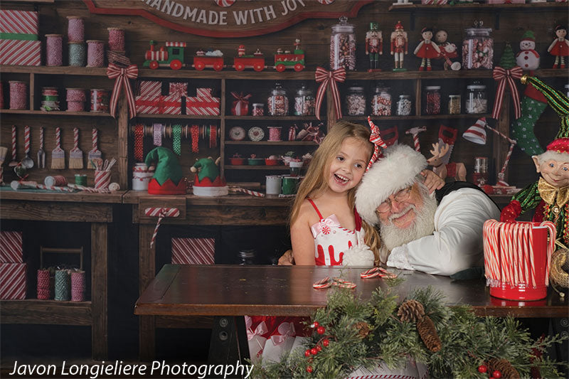 Peppermint Hollow Santa's peppermint workshop photography backdrop shown with a young girl in a candy dress laughing beside Santa at the workshop table, wide view CP-8912