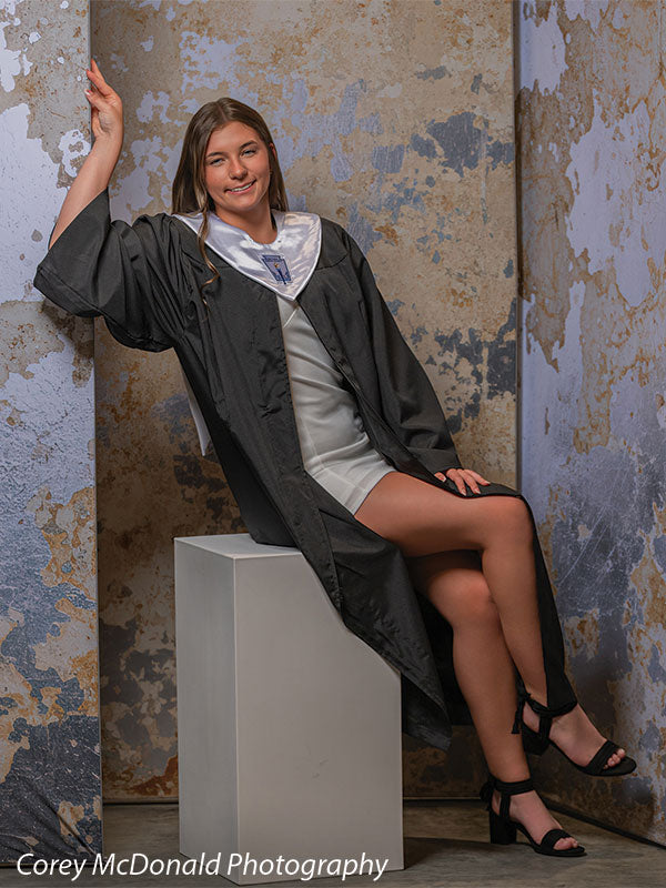 Photography posing cube shown with a senior girl in a black graduation gown seated on the white posing cube with an arm raised in a vertical portrait PC