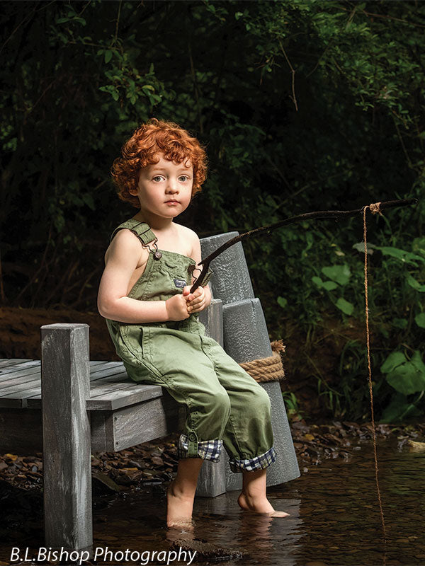 Pier and piling beach prop set shown with a red-haired toddler in green overalls seated beside the gray piling posts holding a twig fishing pole over the creek BEACH-SET