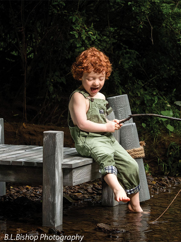 Pier and piling beach prop set shown with a red-haired toddler in green overalls laughing with head tilted down while holding the fishing pole on the pier BEACH-SET