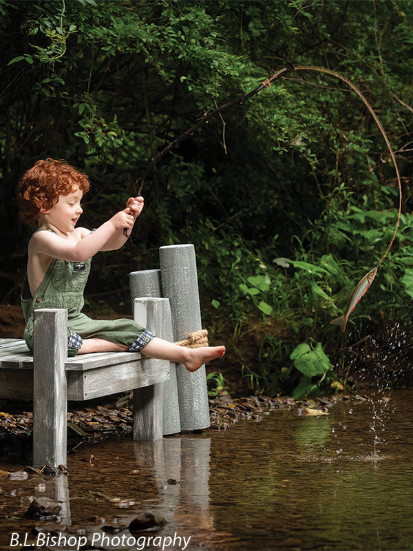 Pier and piling beach prop set shown with a red-haired toddler in green overalls reeling in a small fish on the line with water splashing beside the pier BEACH-SET