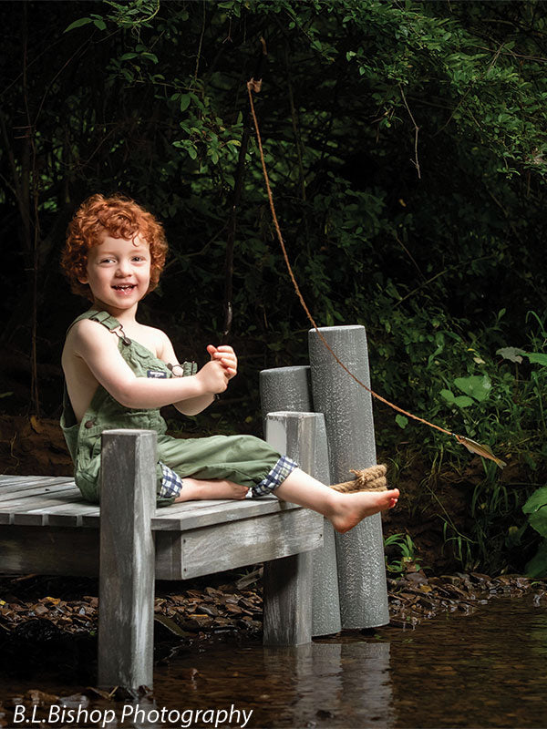 Pier photography prop shown with a red-haired toddler in green overalls grinning on the pier with feet tucked up beside the gray piling posts PIER-3