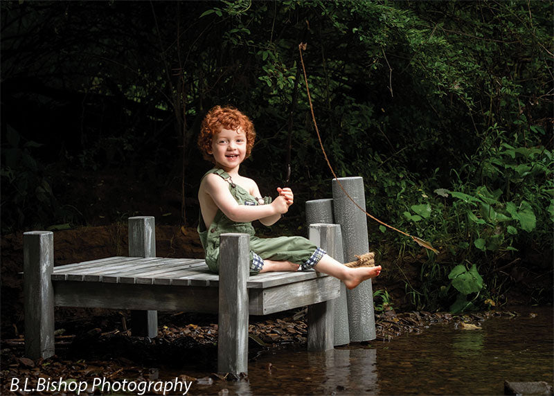 Pier photography prop shown with a red-haired toddler in green overalls smiling on the pier with the fishing pole propped against the gray piling posts PIER-3