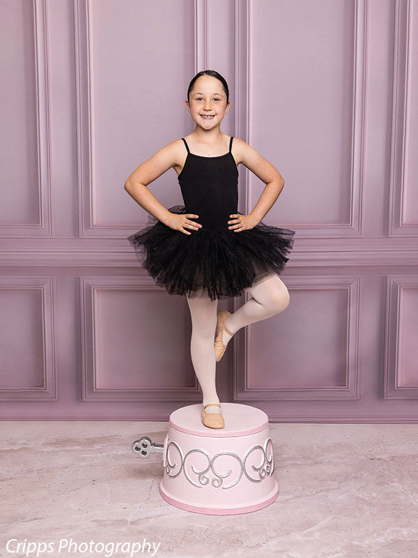 Child dancer poses on a pink and silver wind-up music box prop in a studio with a soft pink paneled wall, ideal for elegant, princess-themed photo shoots.