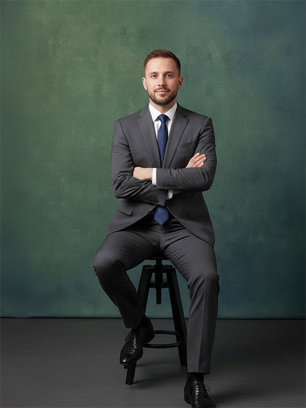 A man wearing a gray suit and blue tie sits on a stool with arms crossed, posed against a rich emerald green textured backdrop with smooth blended tones.
