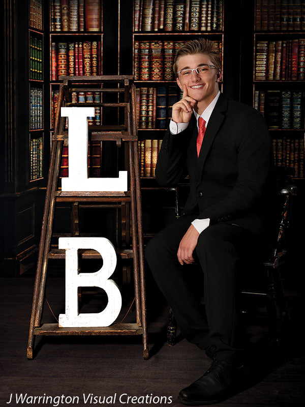 Teen boy in suit seated next to a rustic wooden ladder with large initials, posed over a rich-toned wood studio floordrop ideal for senior or grad photos.