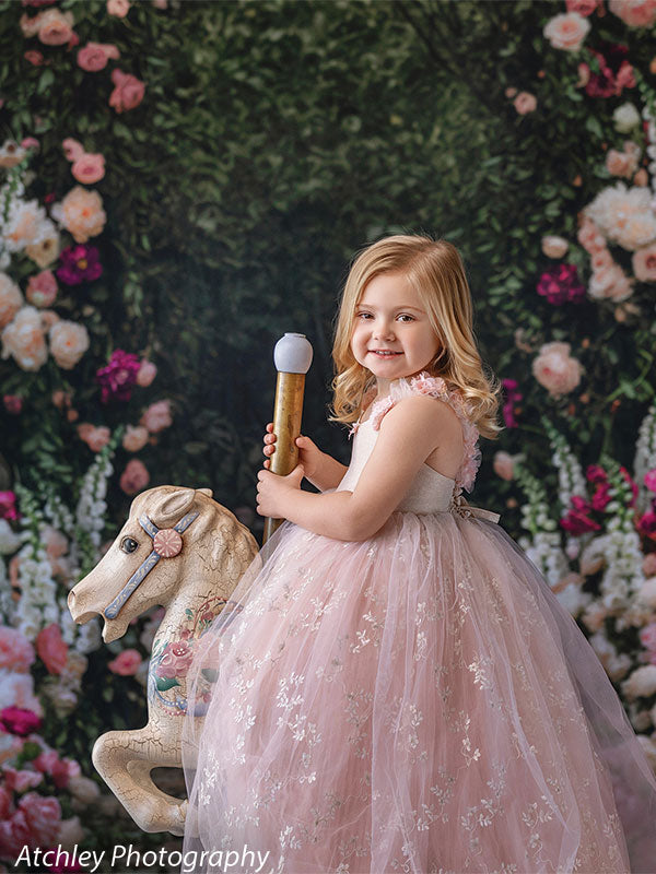 A young girl wearing a blush pink tulle dress smiles while sitting on a white rocking horse, posed in front of a lush garden backdrop with a floral arch, greenery, and blooming roses.
