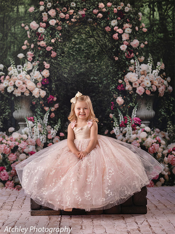 A young girl in a blush pink tulle dress sits centered on a low cushion, smiling toward the camera, posed in front of a romantic floral archway backdrop with roses and greenery.
