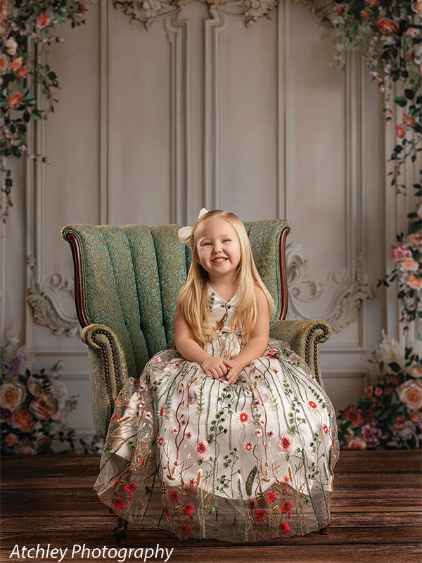 A young girl wearing a floral dress sits centered on a green upholstered chair, smiling toward the camera, posed in front of a romantic vintage wall backdrop framed with flowers.
