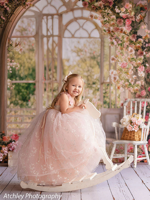A young girl in a blush pink tulle dress sits on a white rocking horse, smiling toward the camera, posed in front of a romantic floral garden room backdrop with arched windows and blossoms.
