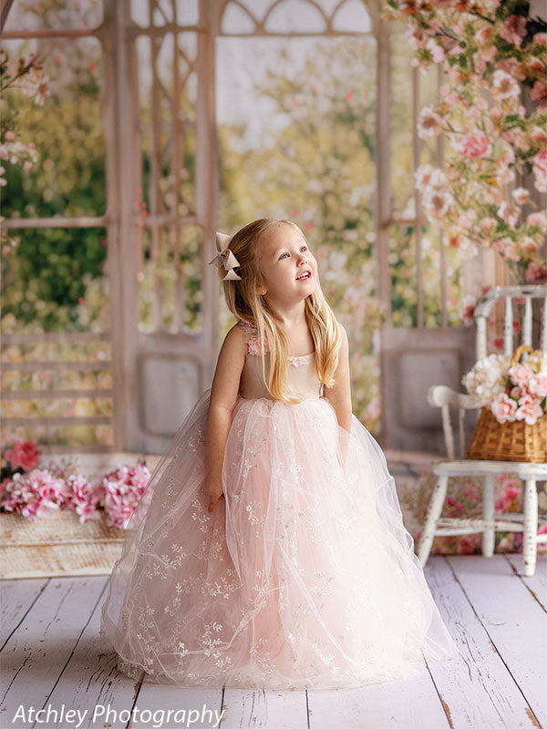 A young girl in a pink tulle dress looks upward with a smile, standing before a romantic garden window backdrop filled with flowering vines and soft natural light.