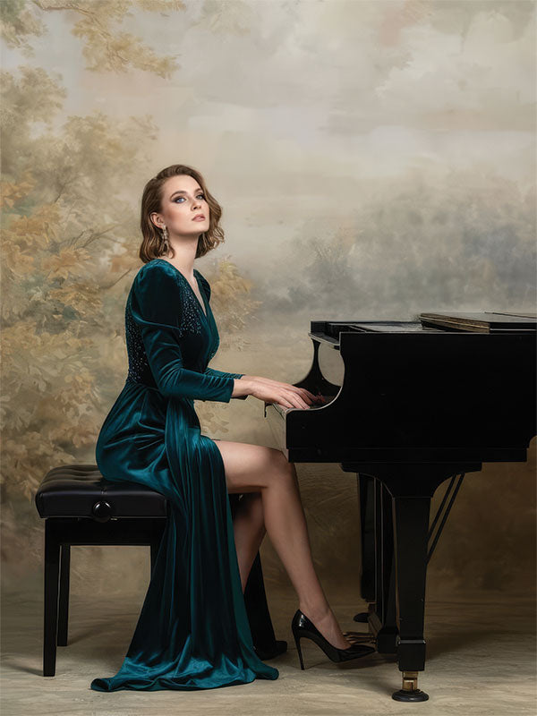 A woman wearing a deep green gown sits at a grand piano, posed against a romantic painted landscape backdrop with soft clouds, muted foliage, and an artistic classical style.
