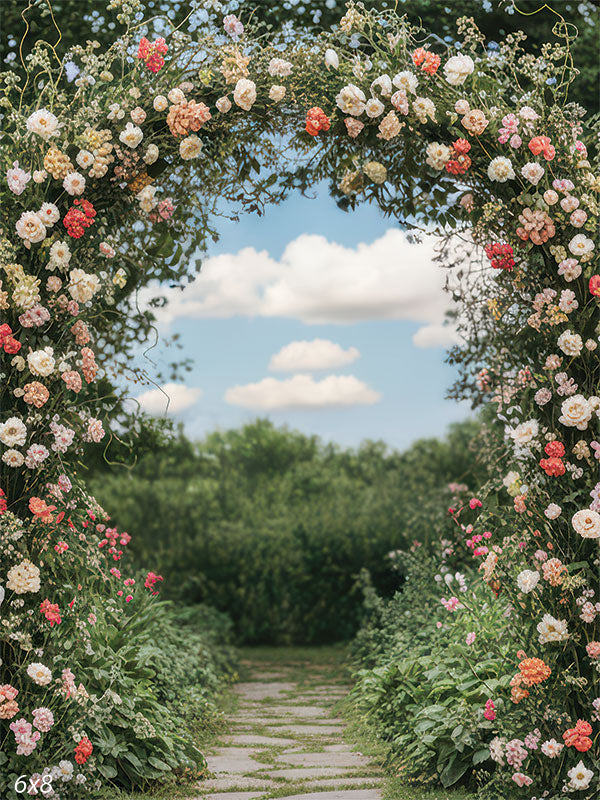 A floral arch bursting with roses in blush and coral tones stands before a blue sky and lush garden path.
